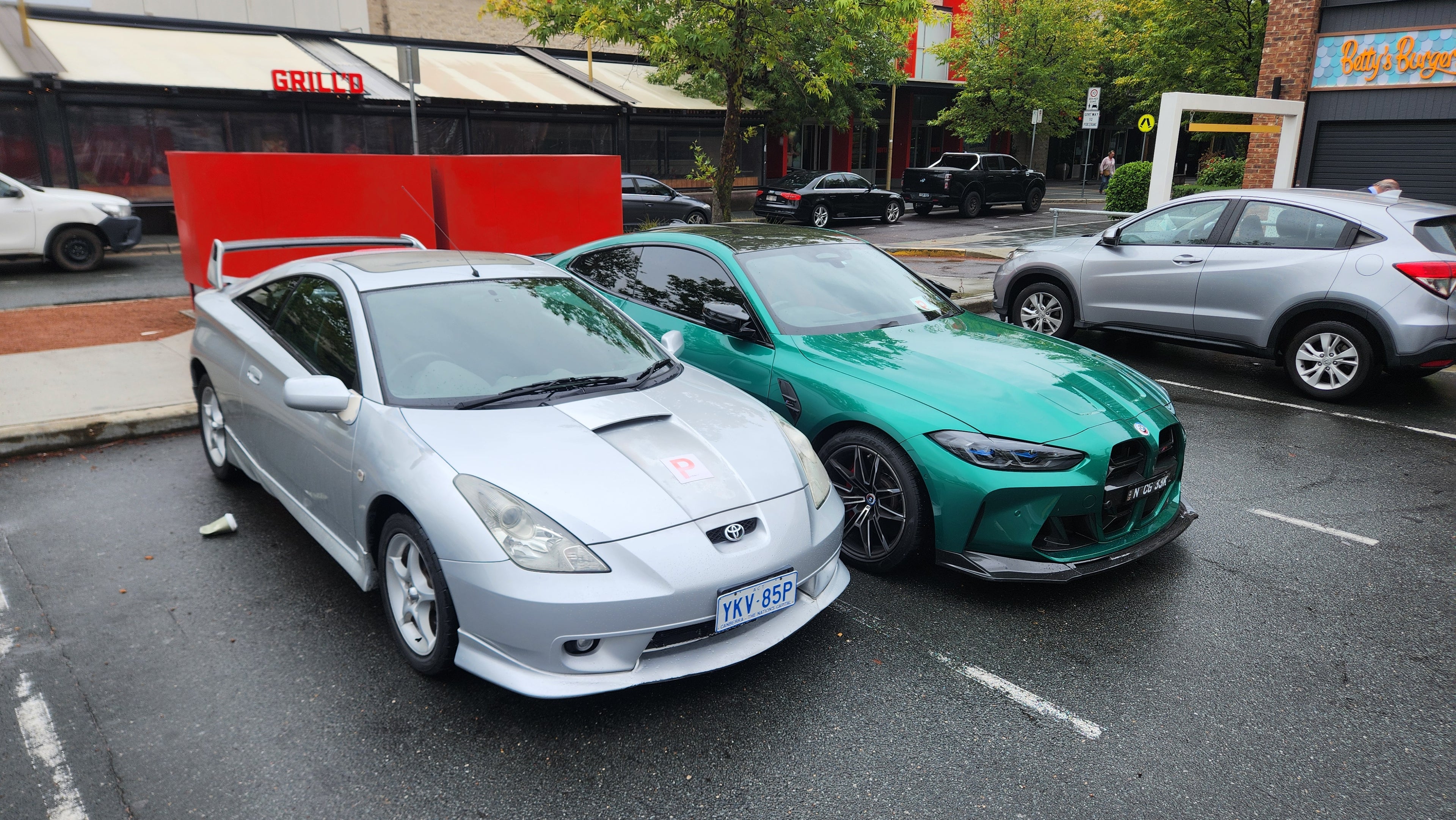 Two cars, a silver Toyota Celica and a green BMW M4 Competition, parked on a street with a building and trees in the background.